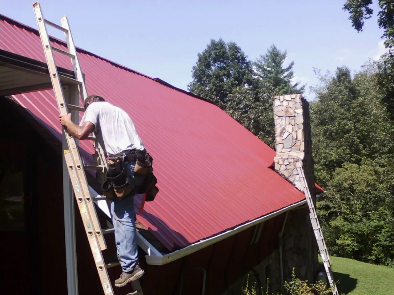 Roofer on a ladder beside a red metal roof and stone chimney — Soffit & Fascia Repair by Roofing Brothers in Okatie